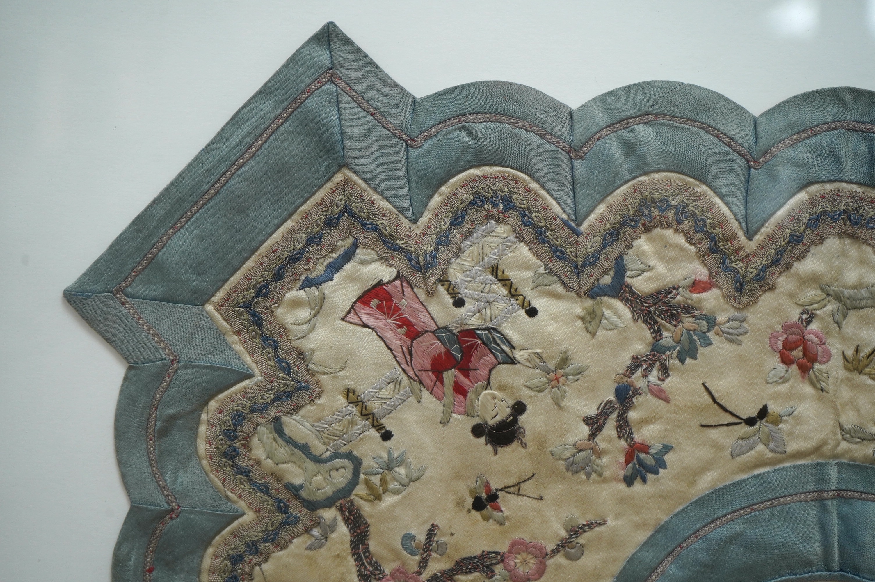 A framed Chinese ornate embroidered collar with scalloped edge, embroidered on cream silk, with polychrome silks, of figures, blossom, butterflies and auspicious symbols, the edges scalloped and braided with an outer blu